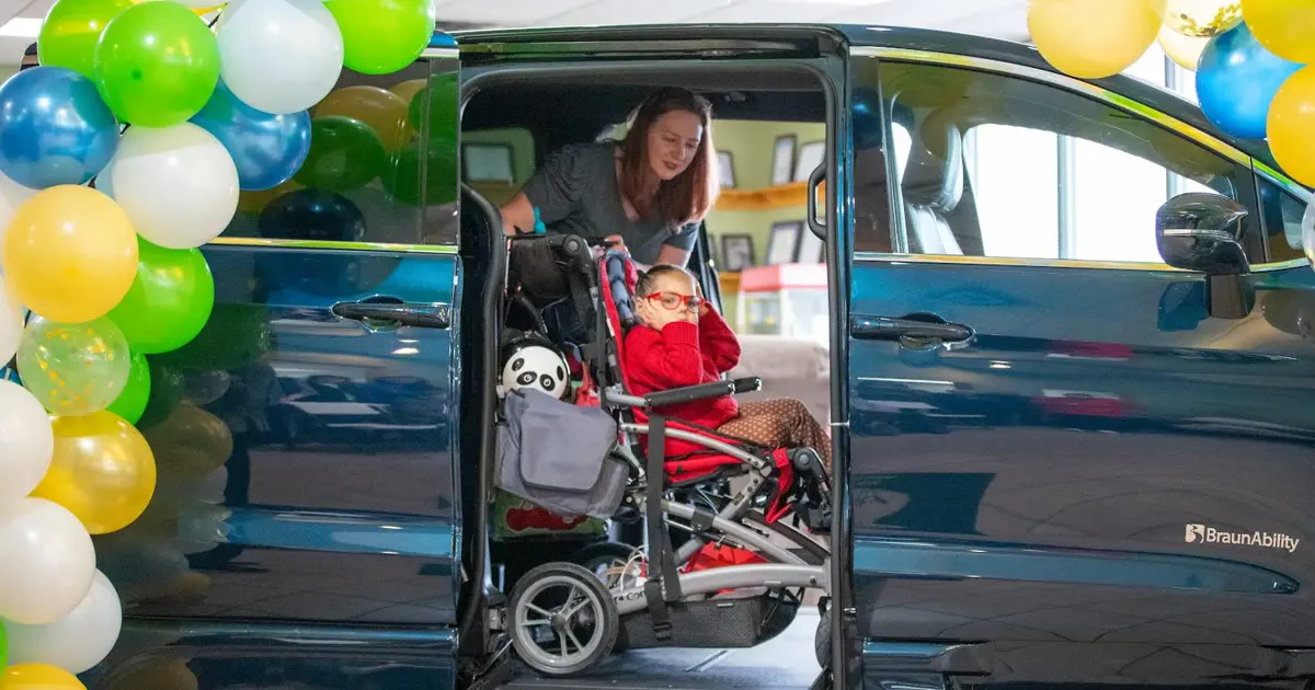 A girl in a red sweater sits in her wheelchair inside an accessible van with its side door open and ramp down, while a woman leans in beside her.